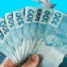 Notas de cem reais em destaque, representando o auxílio de R$ 7.300 liberado pela Caixa para famílias brasileiras.