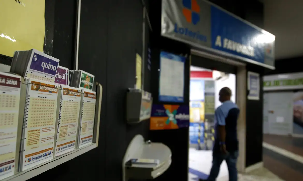 Casa lotérica com cartelas da Quina, Lotomania e Mega-Sena expostas em painel próximo à entrada.