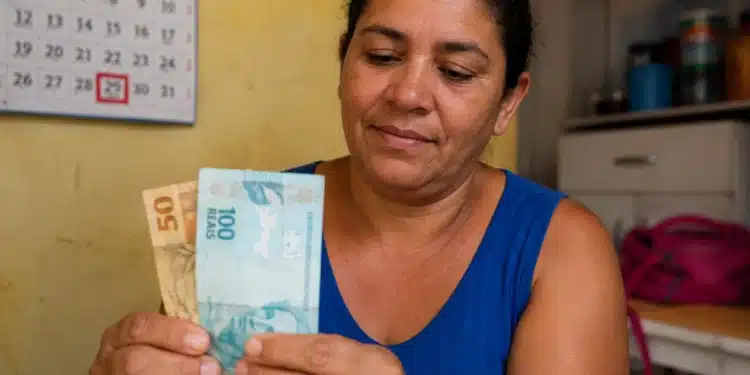 Mulher segurando cédulas de reais em ambiente doméstico, representando o pagamento final de maio do Bolsa Família.