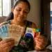 Mulher segurando cartão do Bolsa Família e contando notas de reais em casa, representando o início da nova rodada de pagamentos do programa em maio.