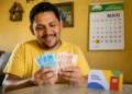Beneficiário do Bolsa Família conta notas de reais em uma casa simples, representando o pagamento para o NIS final 7 em maio, com parcelas extras.