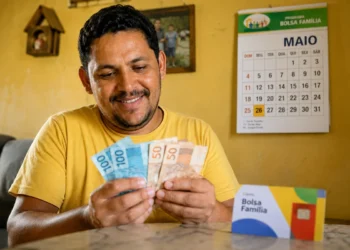 Beneficiário do Bolsa Família conta notas de reais em uma casa simples, representando o pagamento para o NIS final 7 em maio, com parcelas extras.