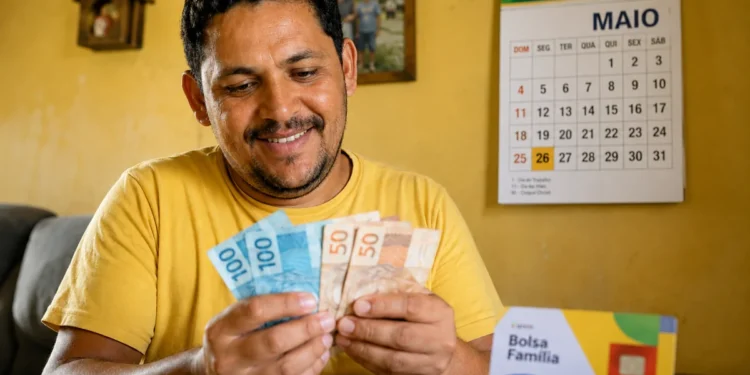 Beneficiário do Bolsa Família conta notas de reais em uma casa simples, representando o pagamento para o NIS final 7 em maio, com parcelas extras.