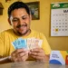 Beneficiário do Bolsa Família conta notas de reais em uma casa simples, representando o pagamento para o NIS final 7 em maio, com parcelas extras.