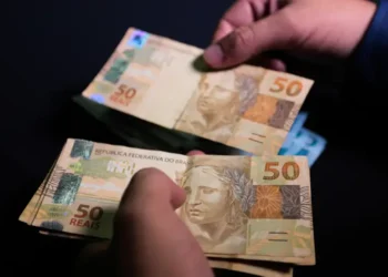 Mãos masculinas segurando cédulas de cinquenta reais brasileiras com fundo escuro, simbolizando recebimento de benefício social