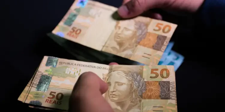 Mãos masculinas segurando cédulas de cinquenta reais brasileiras com fundo escuro, simbolizando recebimento de benefício social