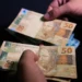 Mãos masculinas segurando cédulas de cinquenta reais brasileiras com fundo escuro, simbolizando recebimento de benefício social