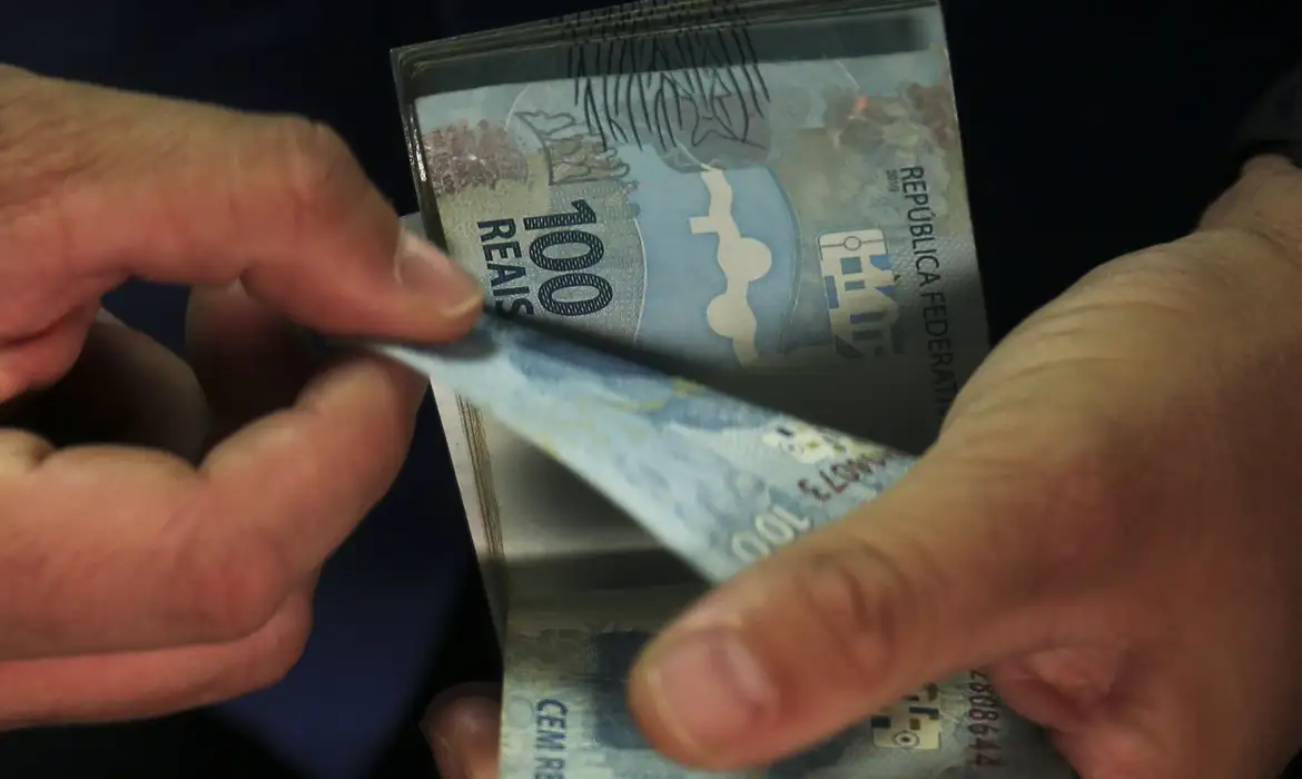 Pessoa contando notas de cem reais durante recebimento do pagamento duplo do INSS