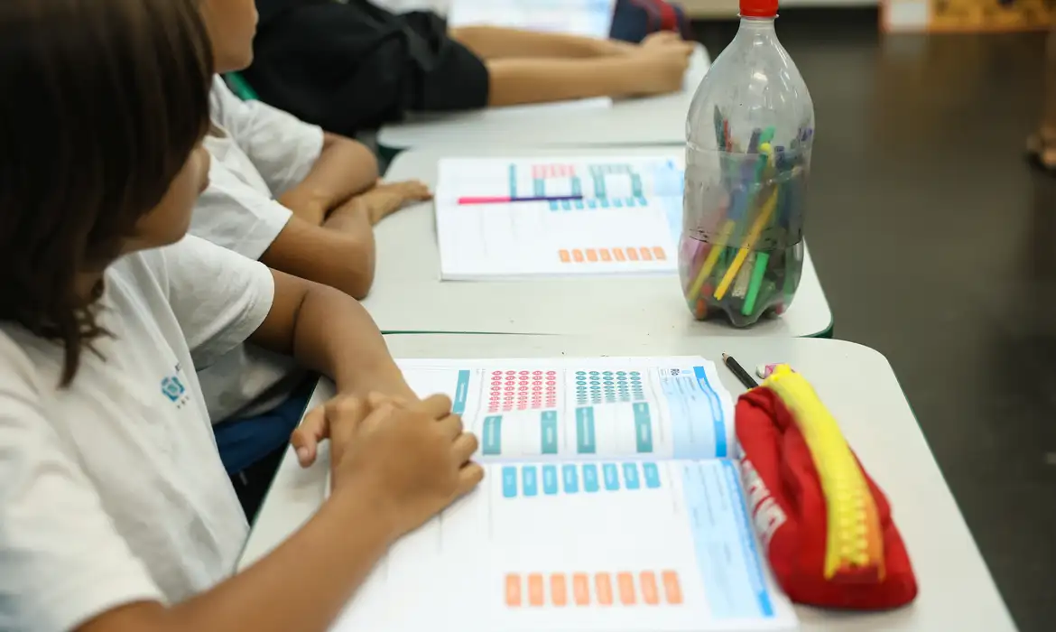 Alunos do ensino fundamental sentados em carteiras escolares preenchem formulários impressos com tabelas coloridas durante atividade em sala de aula
