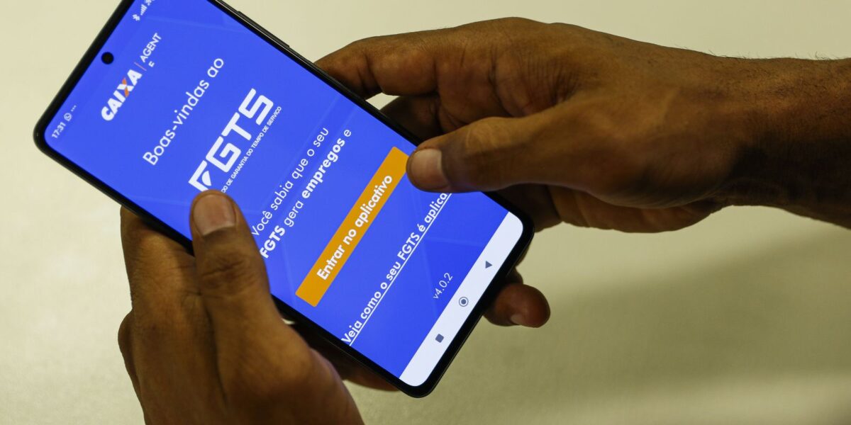 Mãos de homem seguram celular com a tela de boas-vindas do aplicativo FGTS da Caixa aberta exibindo o botão de acesso