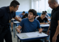 Jovem de camiseta azul sentado em carteira escolar sendo questionado por dois agentes de segurança uniformizados.