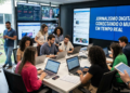 Jornalistas trabalham em redação digital moderna com laptops, telões exibindo redes sociais e painel azul.