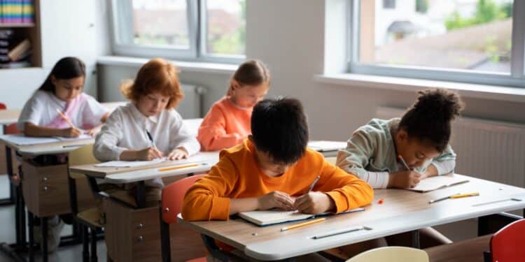 Imagem ilustrativa de cinco crianças escrevendo em cadernos sentadas em carteiras de uma sala de aula, cena usada para tratar do prazo final de envio dos dados das escolas ao Sistema Educacenso no Censo Escolar 2026
