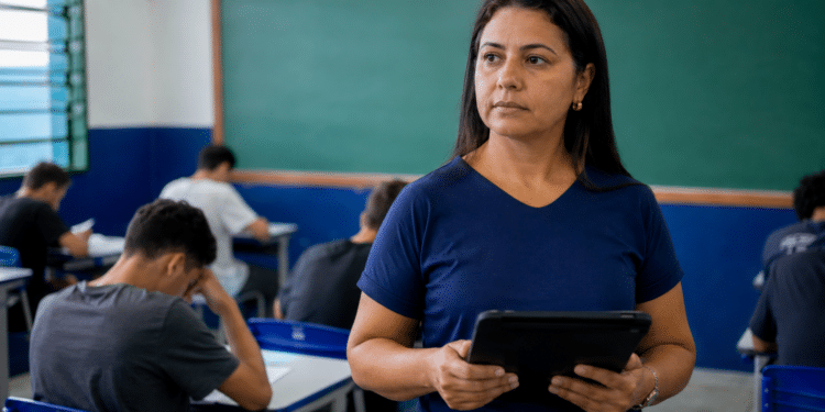 Professora de camiseta azul marinho segura tablet preto enquanto alunos fazem prova em sala com lousa verde.