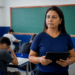 Professora de camiseta azul marinho segura tablet preto enquanto alunos fazem prova em sala com lousa verde.