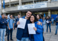 Dois jovens sorriem e exibem documentos oficiais em frente a um centro de formação enquanto outros aprovados comemoram ao fundo