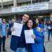 Dois jovens sorriem e exibem documentos oficiais em frente a um centro de formação enquanto outros aprovados comemoram ao fundo