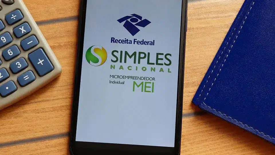 Celular exibe logos da Receita Federal, Simples Nacional e MEI ao lado de calculadora sobre uma mesa de madeira