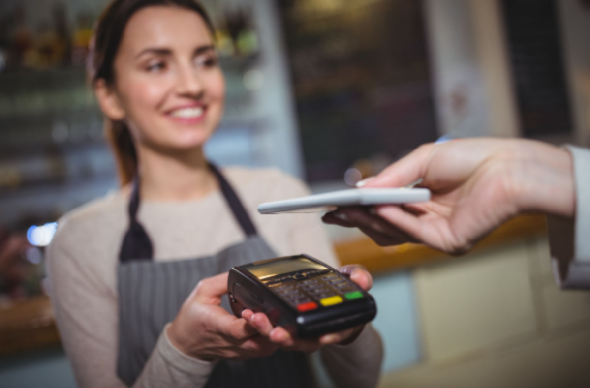Atendente sorridente segura maquininha enquanto cliente aproxima celular para pagamento NFC