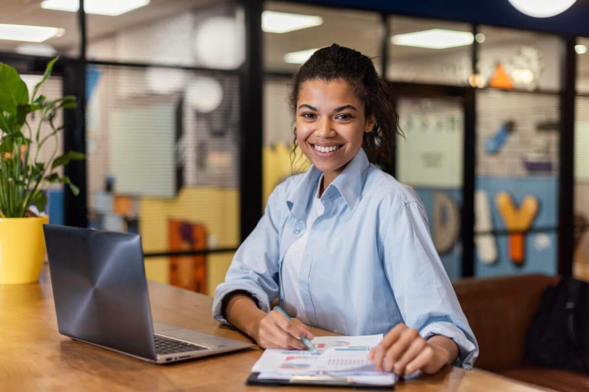 Jovem sorri sentada à mesa com notebook e gráficos impressos, em ambiente de escritório.