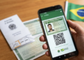 Mão segurando smartphone com app e-Título aberto, exibindo título digital com foto e QR Code, bandeira do Brasil ao fundo