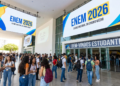 Dezenas de estudantes com mochilas reunidos na entrada de local de prova do ENEM 2026 sob banners azuis e amarelos.