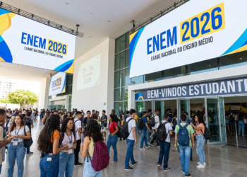 Dezenas de estudantes com mochilas reunidos na entrada de local de prova do ENEM 2026 sob banners azuis e amarelos.
