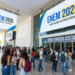 Dezenas de estudantes com mochilas reunidos na entrada de local de prova do ENEM 2026 sob banners azuis e amarelos.