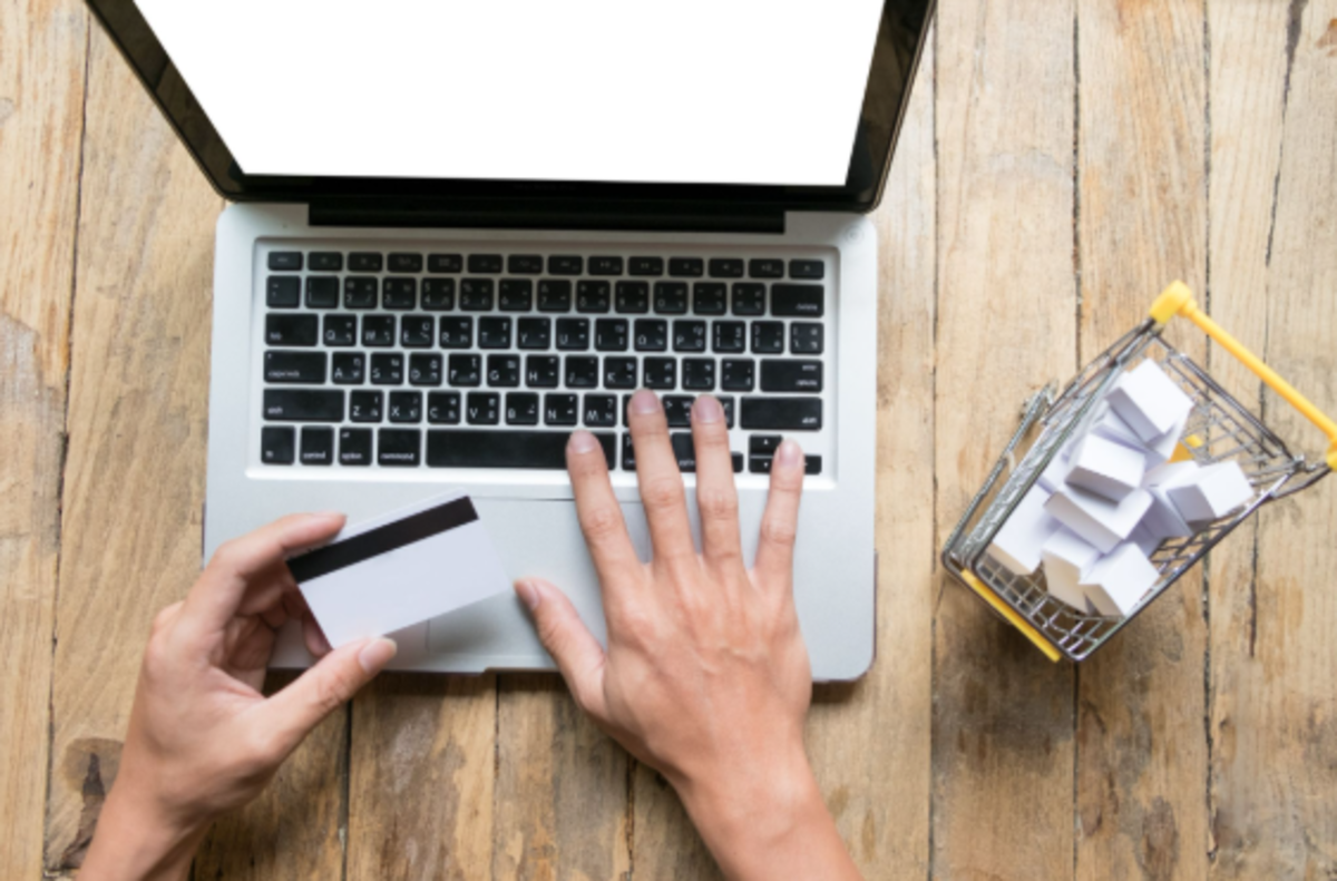 Mãos digitando em laptop prateado segurando cartão bancário, carrinho de compras em miniatura com caixas ao lado
