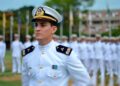 Oficial da Marinha de uniforme branco e quepe em destaque diante de formação militar enfileirada ao fundo em pátio externo
