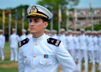 Oficial da Marinha de uniforme branco e quepe em destaque diante de formação militar enfileirada ao fundo em pátio externo