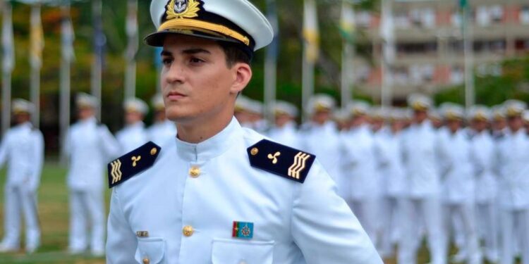 Oficial da Marinha de uniforme branco e quepe em destaque diante de formação militar enfileirada ao fundo em pátio externo