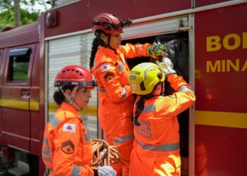 Bombeiras militares de Minas Gerais com uniforme operacional ajustam equipamentos ao lado de viatura