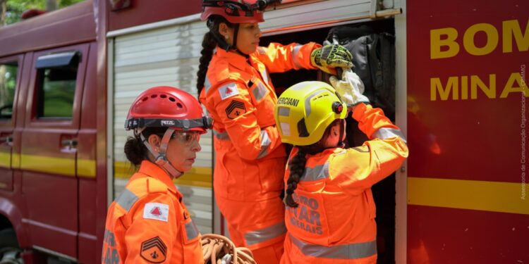 Bombeiras militares de Minas Gerais com uniforme operacional ajustam equipamentos ao lado de viatura
