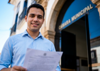 Homem jovem de camisa azul clara segura documento com brasão, sorrindo em frente à Câmara Municipal.