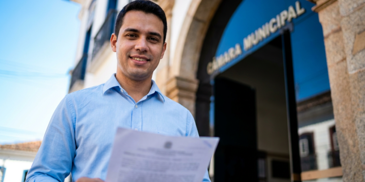 Homem jovem de camisa azul clara segura documento com brasão, sorrindo em frente à Câmara Municipal.