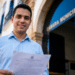 Homem jovem de camisa azul clara segura documento com brasão, sorrindo em frente à Câmara Municipal.