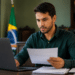 Homem de camisa verde-escura segura folha de papel enquanto olha para laptop em escritório com bandeira brasileira ao fundo.