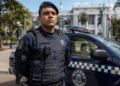 Guarda municipal fardado com colete balístico azul marinho ao lado de viatura com emblema da corporação.