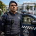 Guarda municipal fardado com colete balístico azul marinho ao lado de viatura com emblema da corporação.