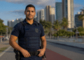 Agente de segurança com colete balístico e uniforme azul-marinho em avenida costeira com palmeiras e arranha-céus.