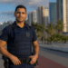 Agente de segurança com colete balístico e uniforme azul-marinho em avenida costeira com palmeiras e arranha-céus.