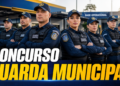 Agentes da Guarda Municipal em formação com coletes e bonés azuis diante de base operacional
