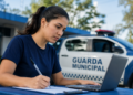 Mulher de camiseta azul marinho escreve em papel enquanto opera laptop cinza ao lado de viatura da Guarda Municipal.