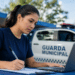 Mulher de camiseta azul marinho escreve em papel enquanto opera laptop cinza ao lado de viatura da Guarda Municipal.