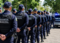 Guardas municipais de costas em fila indiana usando uniforme azul marinho, bonés e coletes, com viatura ao fundo.