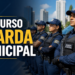 Quatro guardas municipais enfileirados com uniforme azul-marinho, boné e colete tático, viatura ao fundo
