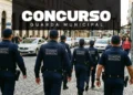 Seis guardas municipais de costas, uniformizados de azul-marinho, caminham em formação por rua de pedestres.