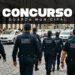 Seis guardas municipais de costas, uniformizados de azul-marinho, caminham em formação por rua de pedestres.