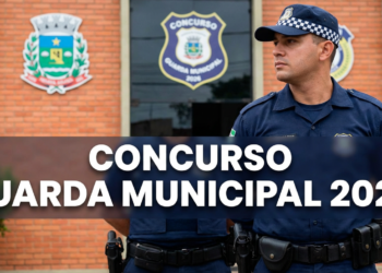Guarda civil uniformizado em frente a prédio público com brasões municipais e emblema de concurso.